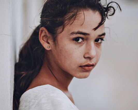 Small gold stud earrings on a real model in everyday/desk context, warm natural light, minimal styling