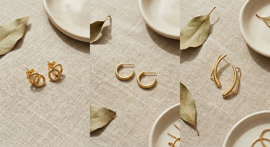 Flat lay of three minimalist gold vermeil earrings: studs, small hoops, and ear climbers on warm neutral stone